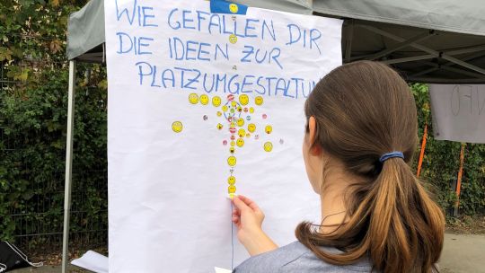 Ein Mädchen klebt Sticker auf ein Plakat mit der Überschrift "Wie gefallen dir die Ideen zu Platzumgestaltung?"