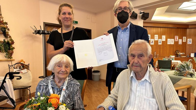 Ein älteres Paar sitzt vor Bezirksvorsteherin Christine Dubravac-Widholm und einem Mann mit Urkunde