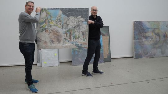 Georg Papai und Gerhard Grimm halte ein Gemälde zwischen sich in die Höhe; sie stehen in einem leeren Saal, Bilder lehnen an der Wand