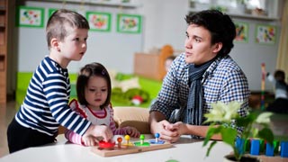 Kindergartenpdagoge sitzt an einem Tisch mit zwei spielenden Kindern.