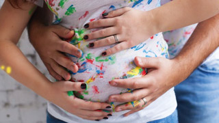 Blick auf den Bauch einer schwangeren Frau, ihre Hnde und die eines Mannes liegen auf dem Bauch, auf dem T-Shirt sind Farbkleckse.