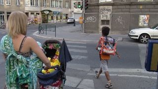 Eine Frau mit Kinderwagen an einer Kreuzung, davor ein Schulkind, das die Strae berquert