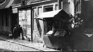 Historische Aufnahme in Schwarz-Wei: Holzkonstruktion mit Ablage, Rckwand und Dach, textilbespannt, mit Verkaufsware. Zwei Personen mit Kind und Hund