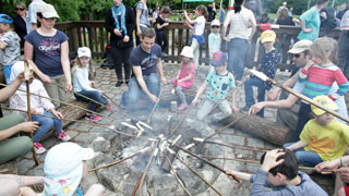 Mehrere Kinder grillen gemeinsam mit ihren Eltern an einer Feuerstelle Wrstchen