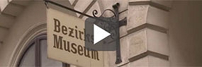 Videoausschnitt mit Play-Button: Eingangsschild mit der Aufschrift "Bezirksmuseum"