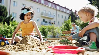 Dijete sa smeđim loknama igra se s plavokosim djetetom u pijesku, u pozadini se može vidjeti stambeno naselje