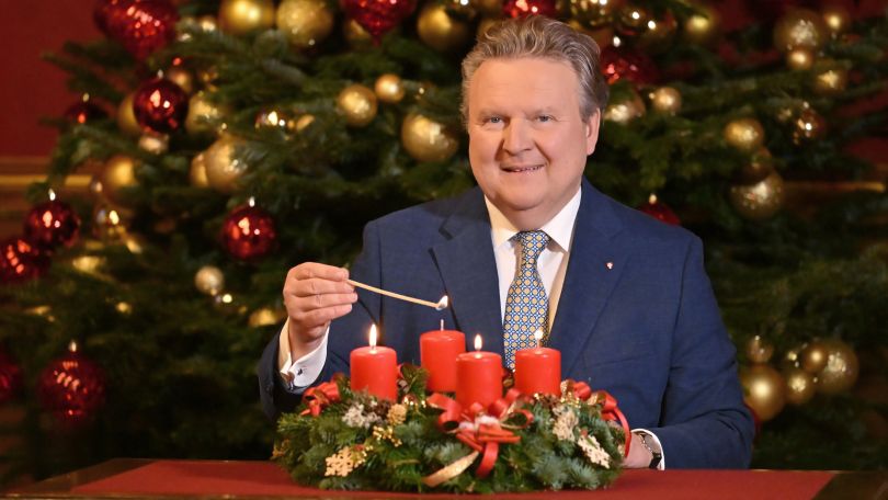 Bürgermeister Ludwig zündet Kerze am Adventkranz an, vor Weihnachtsbaum sitzend.