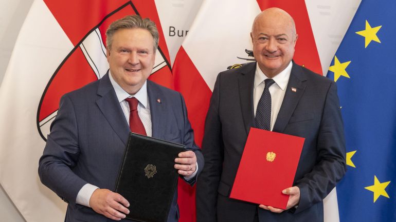 Bürgermeister Michael Ludwig und Bundeskanzler Christian Stocker stehen vor den Fahnen Wiens, Österreichs und der EU und halten jeweils eine Mappe mit dem Wiener Landeswappen bzw. österreichischen Bundeswappen in ihren Händen