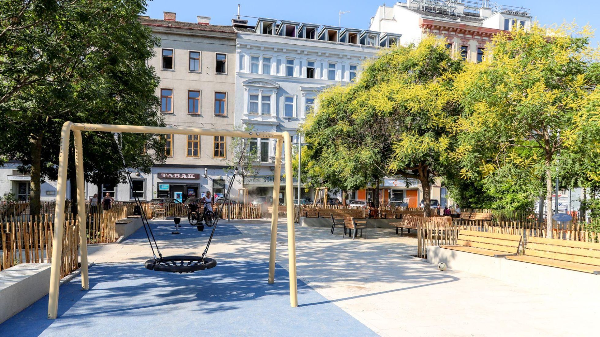 copyright: Stadt Wien/Christian Fürthner Nestschaukel auf heller Pflasterung, daneben Sitzgelegenheiten und blühende Bäume.
