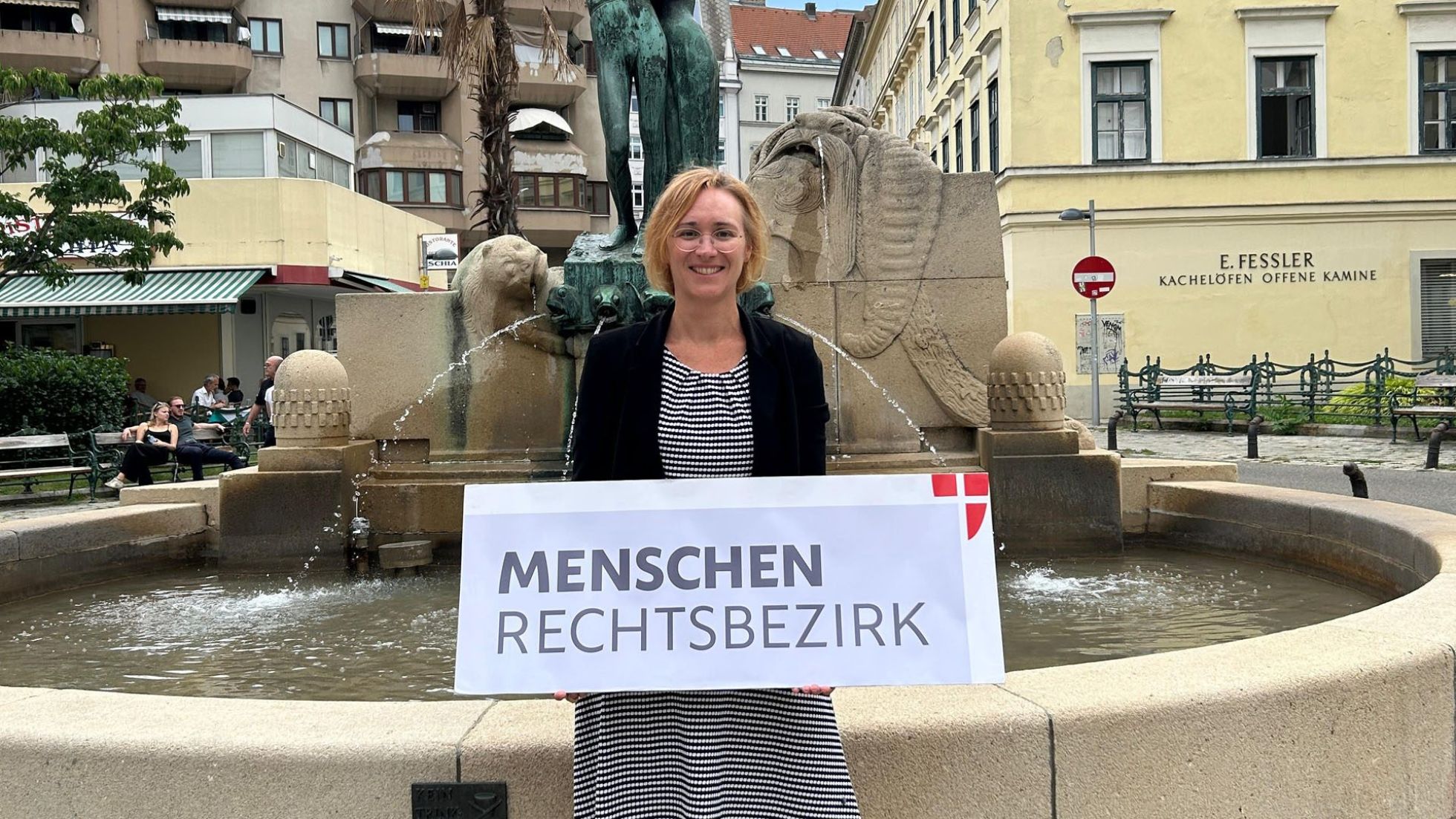Lea Halbwidl steht vor einem Brunnen und hält ein schild mit der Aufschrift "MEnschenrechtsbezirk" in den Händen