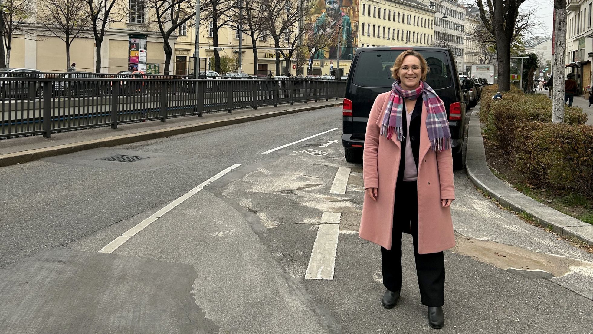 Eine Frau in rosa Wintermantel steht auf einer Straße mit Fahrbahn, Mehrzweckstreifen zum Radfahren und Parkplätzen.