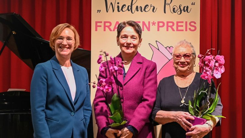3 Frauen, 2 davon halten rosa Blumen in der Hand.