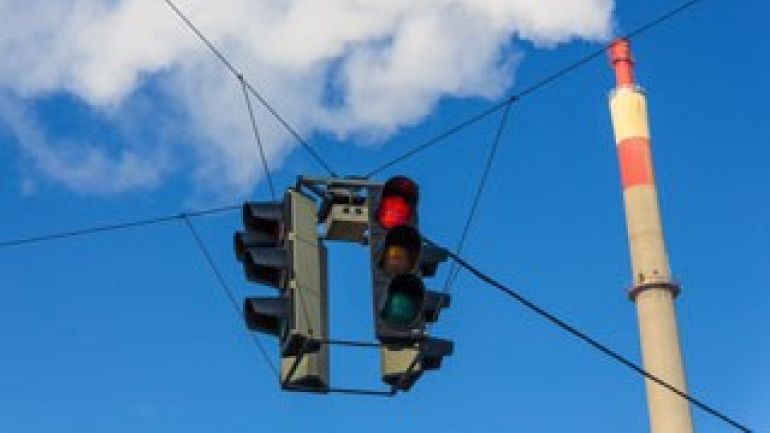 Schlot eines Industriebetriebes, aus dem weißer Rauch austritt, und eine rote Ampel