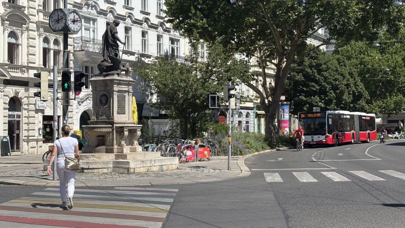Eine Fußgängerin, ein Fahrradfahrer und ein Bus am Margaretenplatz. Man sieht auch Bäume und eine Statue.