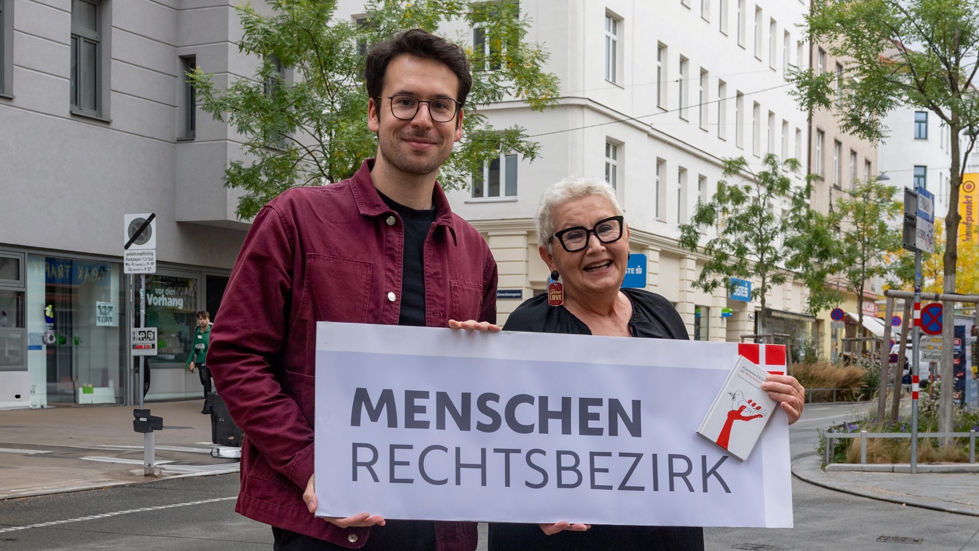 Bezirksvorsteher Michael Luxenberger hält mit einer Person Plakat mit Aufschrift "Menschenrechtsbezirk" in der Hand