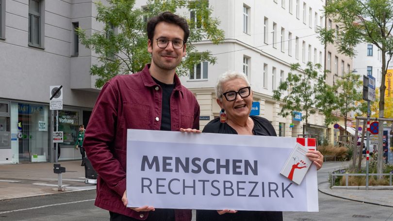Bezirksvorsteher Michael Luxenberger hält mit einer Person Plakat mit Aufschrift "Menschenrechtsbezirk" in der Hand
