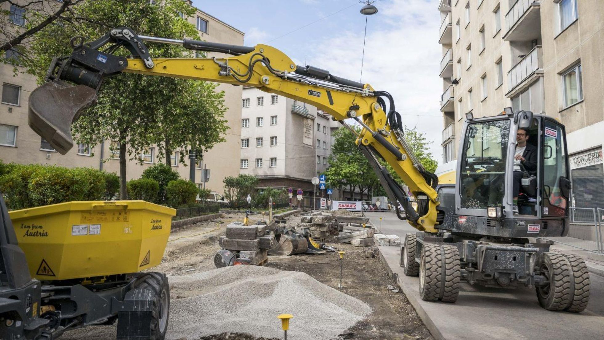 copyright: Stadt Wien/Karo Pernegger Ein Bagger gräbt eine Straße auf, daneben Wohnbauten, Bäume und Sträucher.
