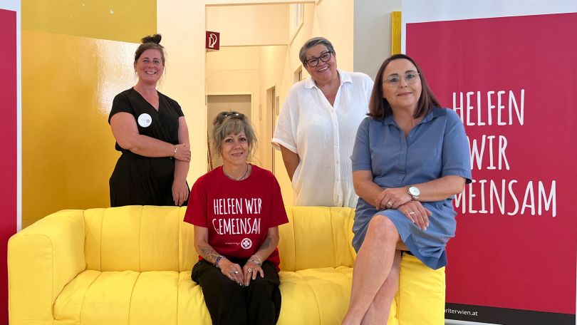 copyright: BV 6 4 Frauen sitzen am beziehungsweise stehen um ein gelbes Sofa; im Hintergrund ein Plakat mit der Aufschrift "Helfen wir gemeinsam"