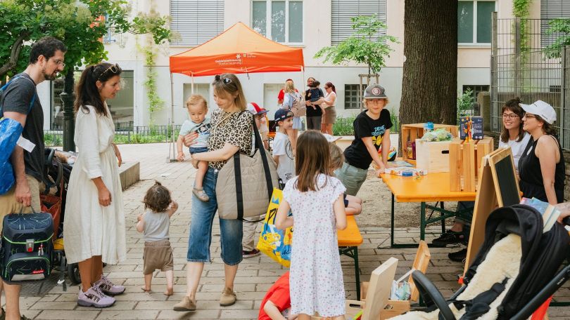 copyright: GB* - Daniel Dutkowski Erwachsene und Kinder beim Spielen und Reden auf einem gepflasterten Platz zwischen Bäumen.