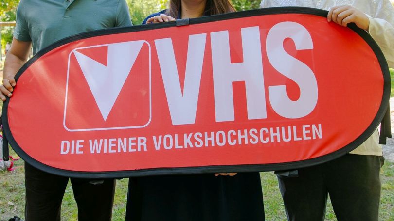 Hände von 3 Personen halten Schild mit Aufschrift "VHS - Volkshochschulen"