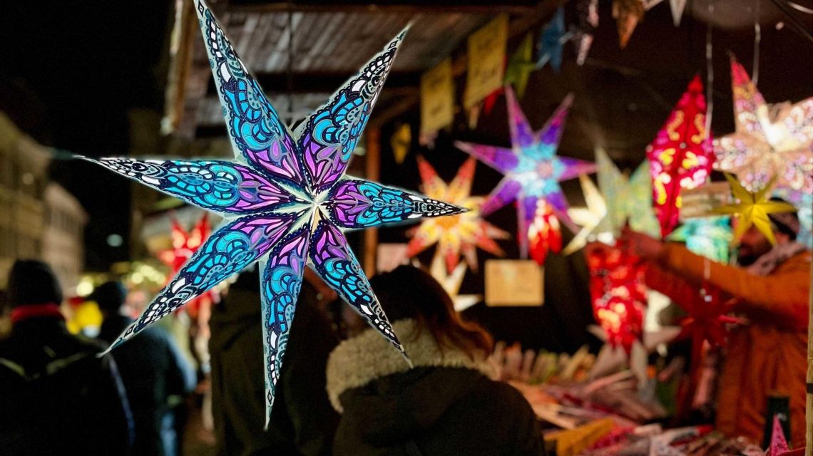 Sternförmige Lampions; im Hintergrund sind verschwommen Menschen erkennbar die den Weihnachtsmarkt besuchen