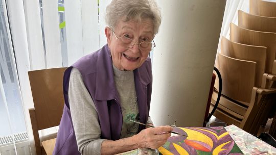 Eine ältere Dame malt mit Pinsel an einem bunten Bild