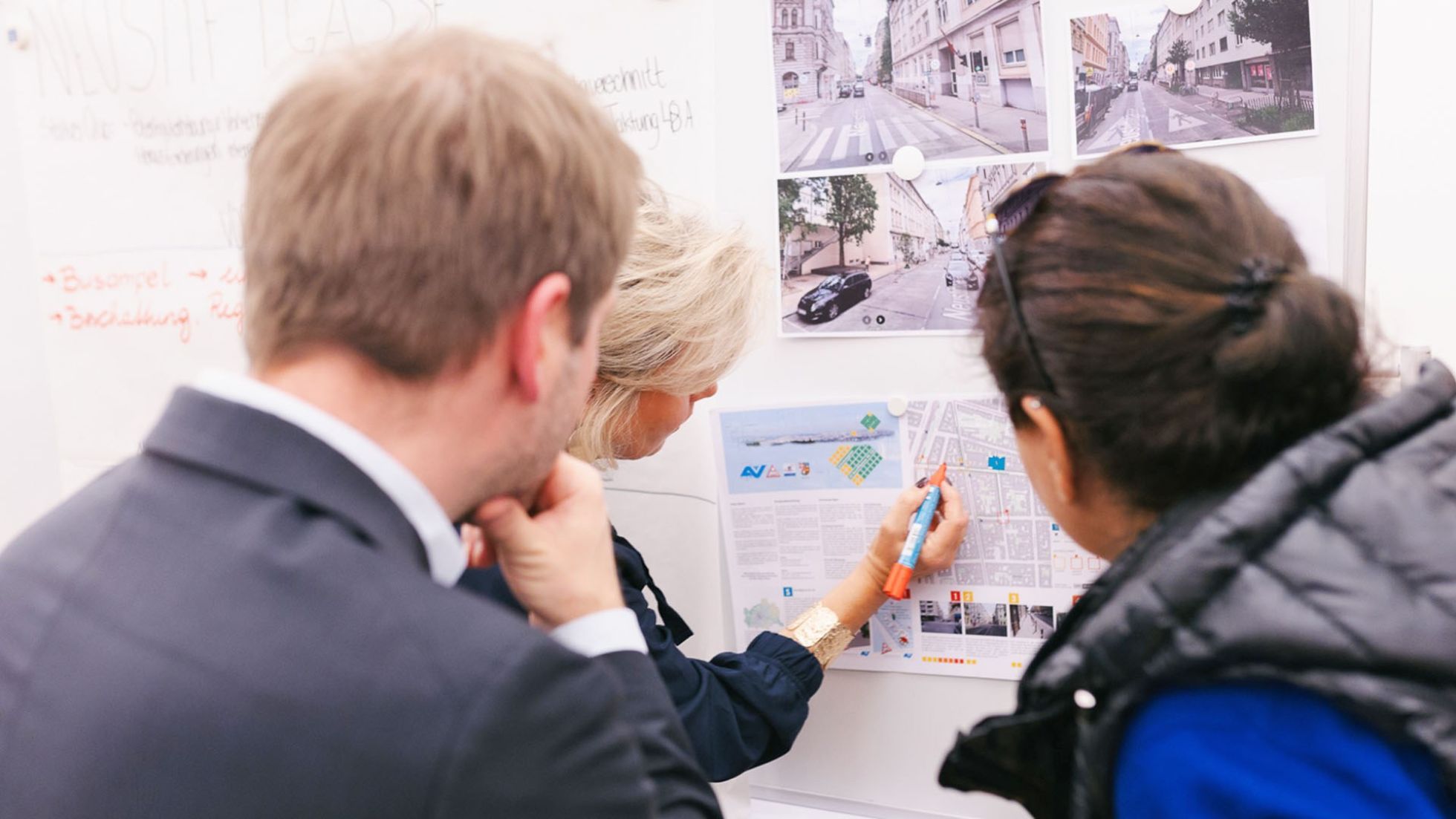 Ein Mann und zwei Frauen stehen vor einer Infotafel mit Fotos von Schulvorplätzen