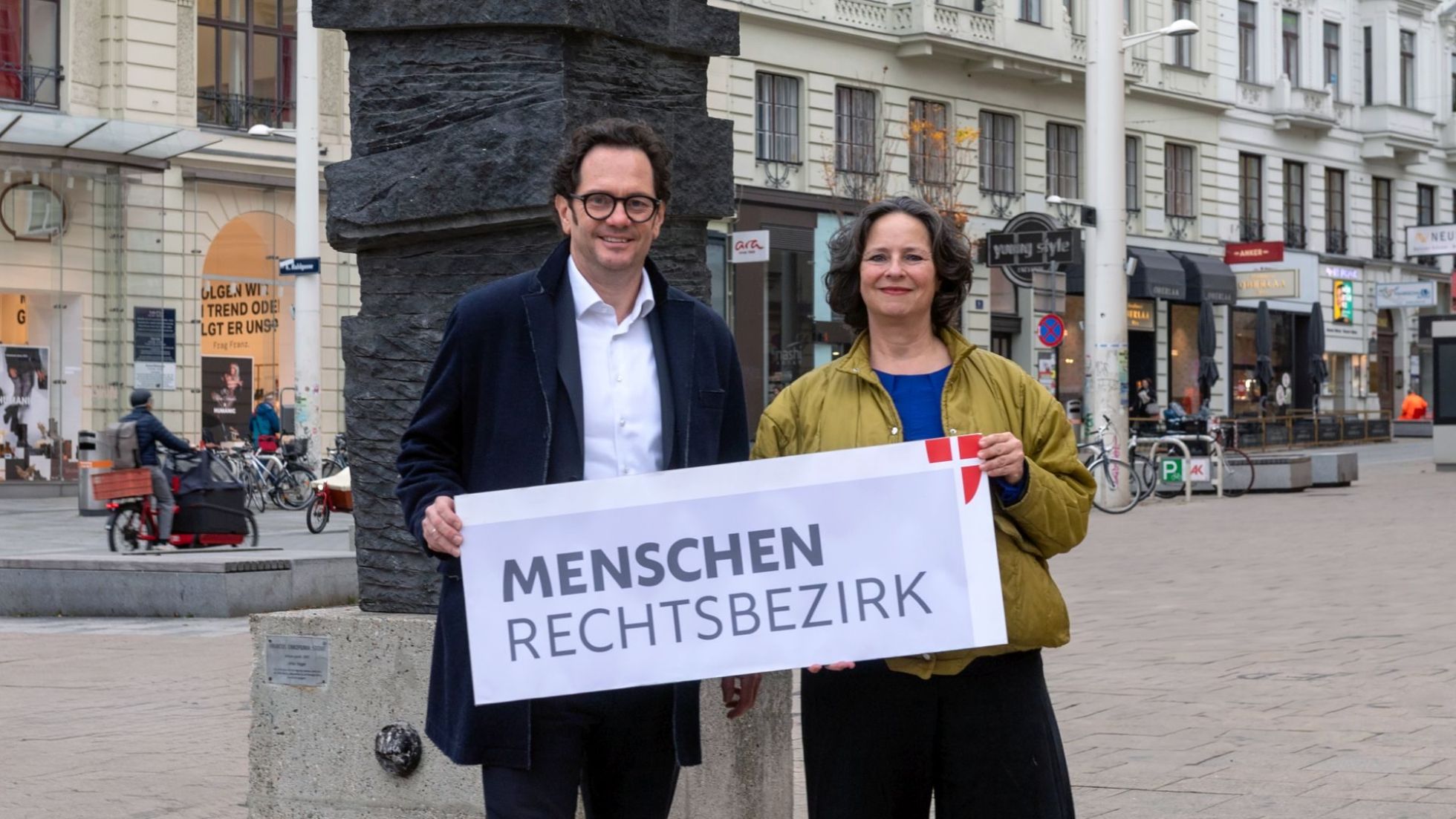 Ein Politiker und eine Politikerin halten ein Schild "Menschenrechtsbezirk", dahinter eine Einkaufsstraße.