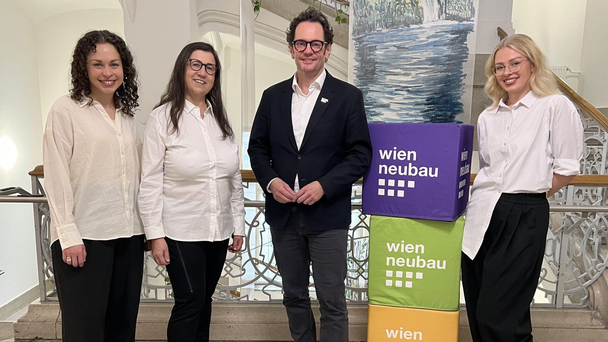 3 Frauen und ein Mann stehen am Gang eines Altbaugebäudes neben Aufstellerwürfel mit der Aufschrift "wien neubau"