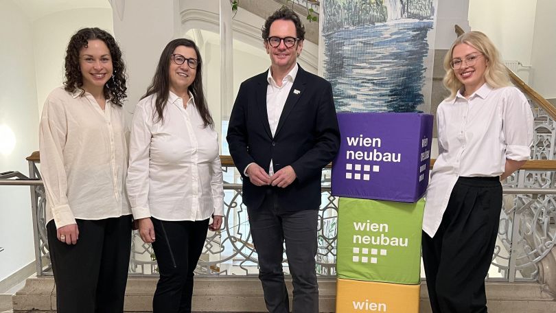 3 Frauen und ein Mann stehen am Gang eines Altbaugebäudes neben Aufstellerwürfel mit der Aufschrift "wien neubau"