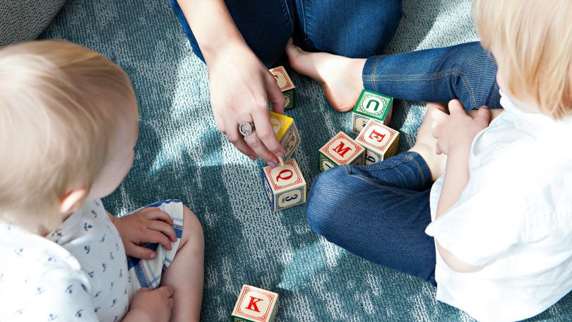 2 Kleinkinder und eine erwachsene Person spielen mit Bauklötzen am Boden