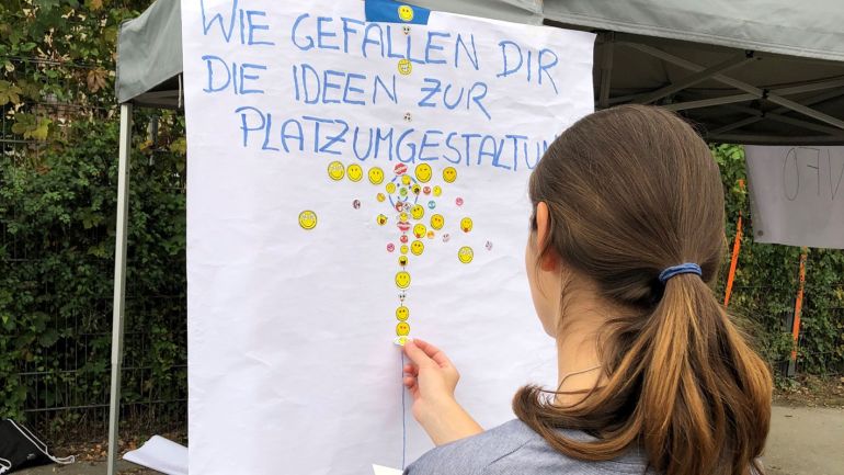 copyright: PlanSinn Ein Mädchen klebt Sticker auf ein Plakat mit der Überschrift "Wie gefallen dir die Ideen zu Platzumgestaltung?"