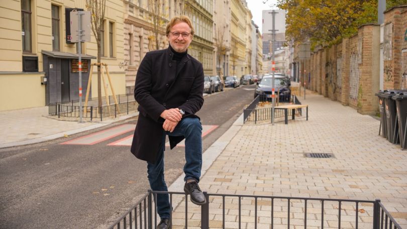 Ein Mann im schwarzen Mantel lehnt auf einem schwarzen Geländer, abgestützt auf ein Knie. Im Hintergrund eine umgestaltete Gasse mit heller Pflasterung.