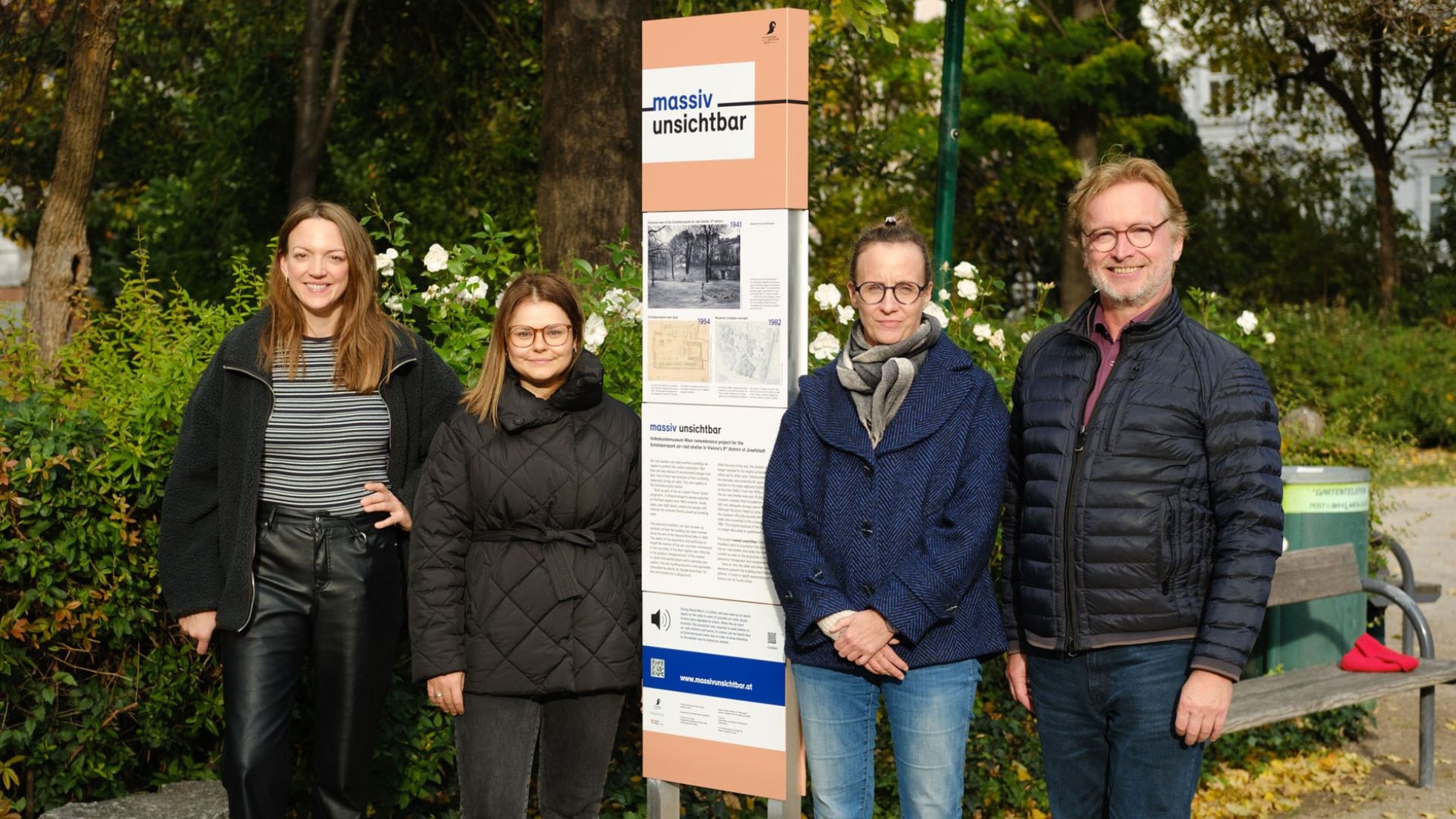 Drei Frauen und ein Mann stehen neben einer Tafel in einem Park.