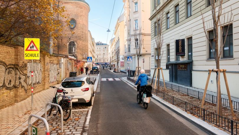 Ein Fahrradfahrer fährt eine schmale Straße zwischen Gebäuden entlang. Rechts parken Autos neben einem breiten Gehweg. Es sind Fahrradständer und ein Zebrastreifen zu sehen.