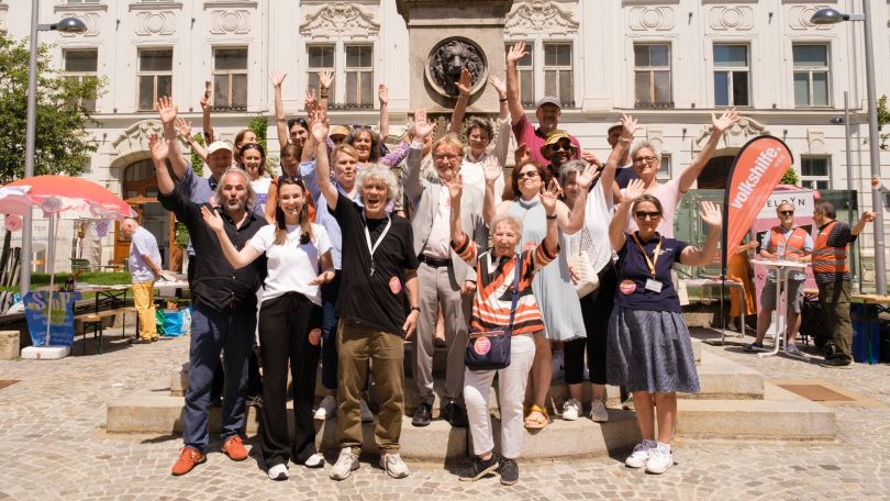 Eine Gruppe Menschen steht vor einem Denkmal und winkt in die Kamera
