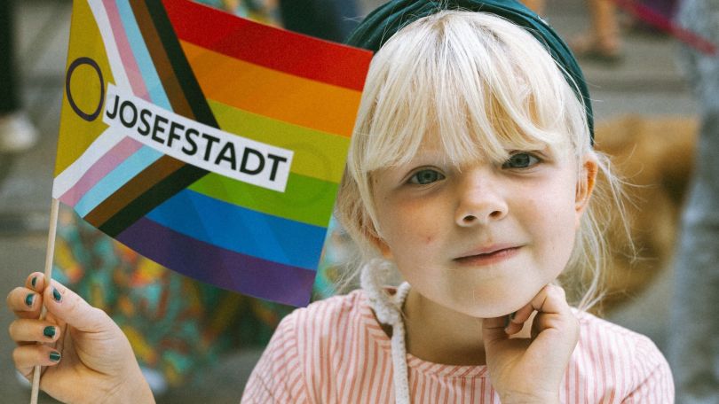 Ein Kind hält eine Regenbogenflage mit der Aufschrift "Josefstadt" in der Hand.