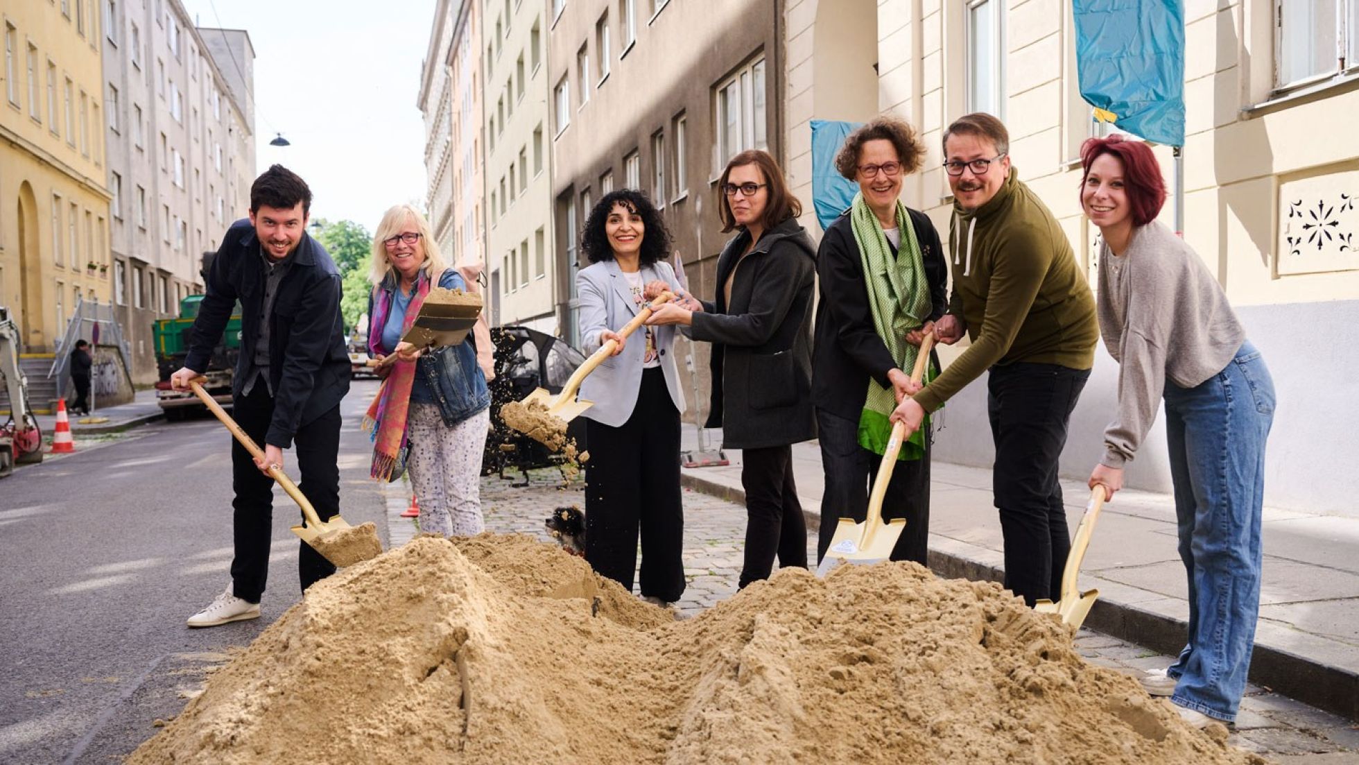 Eine Gruppe von Frauen und Männern steht vor einem Sandhaufen; sie halten Spaten in den Händen