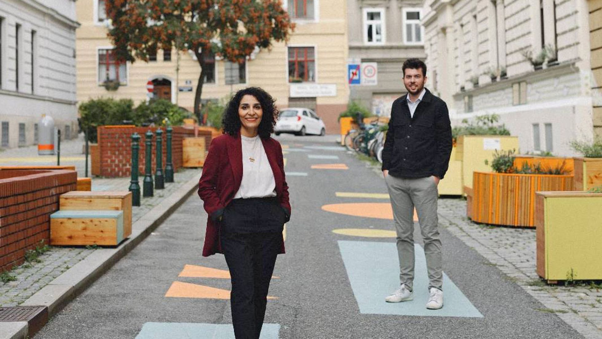 Eine Frau und ein Mann stehen auf bunten Feldern in der Mitte einer Straße. Links und rechts sind Sitzmöglichkeiten und Hochbeete aus Holz zu sehen.