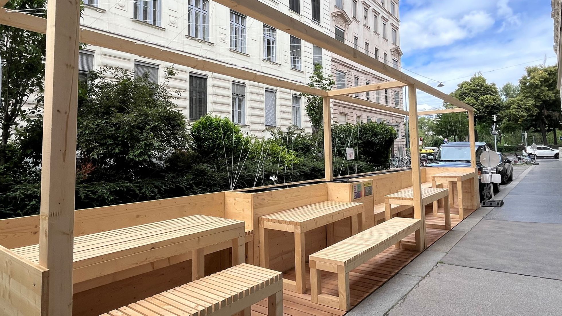 copyright: BV 9 In der Parkspur stehen Bänke und Tische aus hellem Holz, die auf 3 Seiten von einem Holzzaun umgeben sind. Straßenseitig ist der Zaun begrünt.
