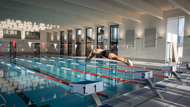 Ein Mann springt von einem Sprungturm in ein Sportbecken in einem Hallenbad.