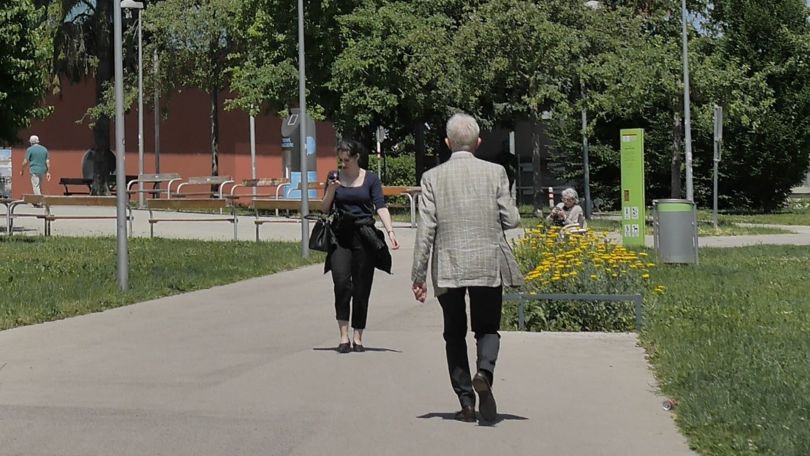 1 Frau und 1 Mann gehen auf betonierten Wegen durch einen Park, 1 Frau sitzt auf einer Bank.