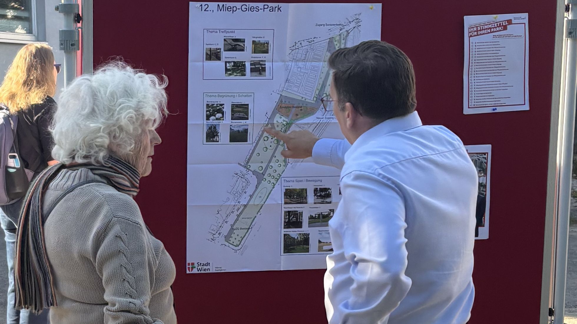 Bezirksvorstehe Wilfried Zankl mit einer älteren Frau vor einer Schautafel mit einem Plan