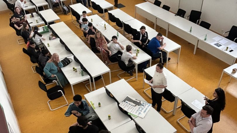 Ein Sitzungssaal von oben. Jugendliche sitzen an den Tischen, eine Person trägt vor. 2 Erwachsene hören zu.