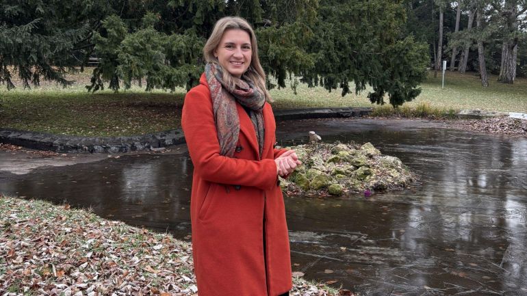 copyright: BV 13 Frau im orangen Mantel vor Teich, dahinter Nadelbäume.