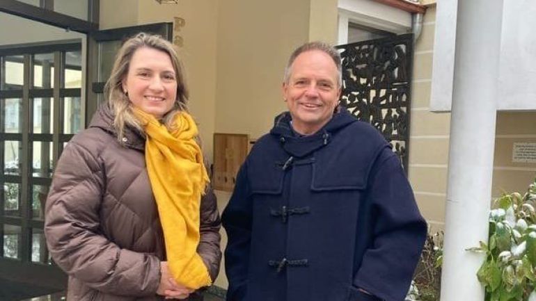 copyright: BV 13 Frau in braunem Wintermantel und gelbem Schal neben einem Mann mit dunkelblauem Wintermantel vor einem Eingang.