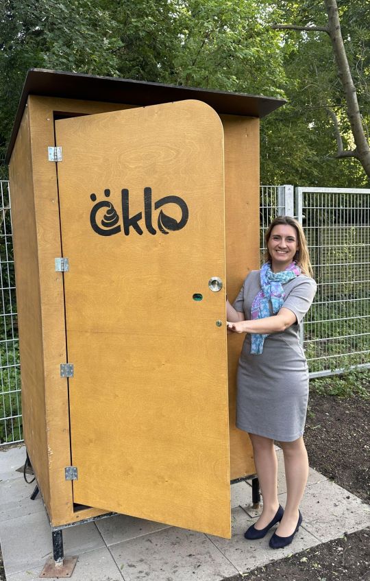 copyright: BV 13 Frau mit blonden Haaren und grauem Rock steht neben öKlo WC-Anlage vor Park
