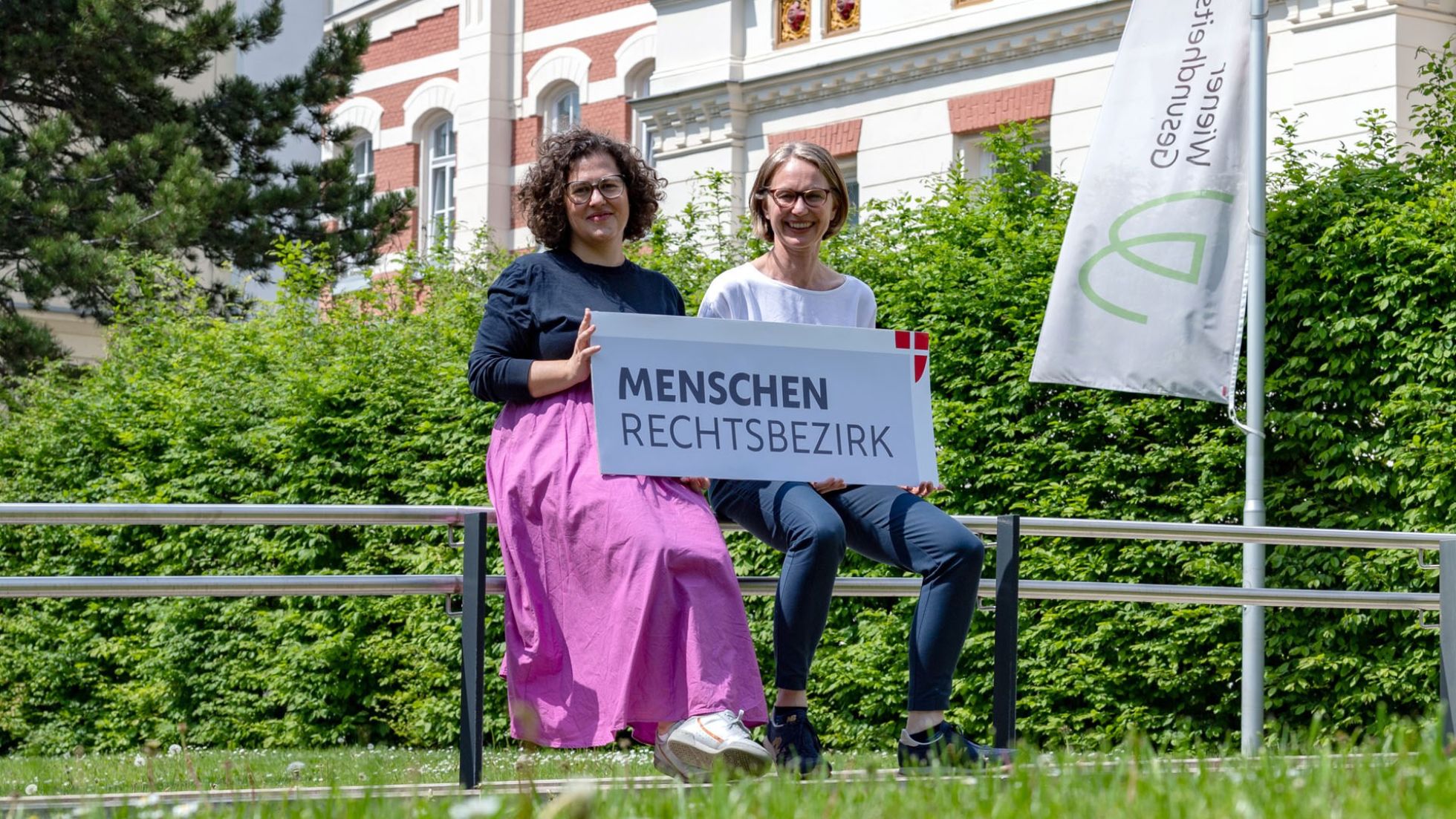 2 Frauen im Grünen halten ein Schild mit der Aufschrift "Menschenrechtsbezirks"