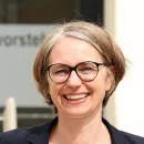 Eine Frau mit Brille lächelt in die Kamera. Sie steht bei Sonnenschein vor dem Eingang zur Bezirksvorstehung Penzing.