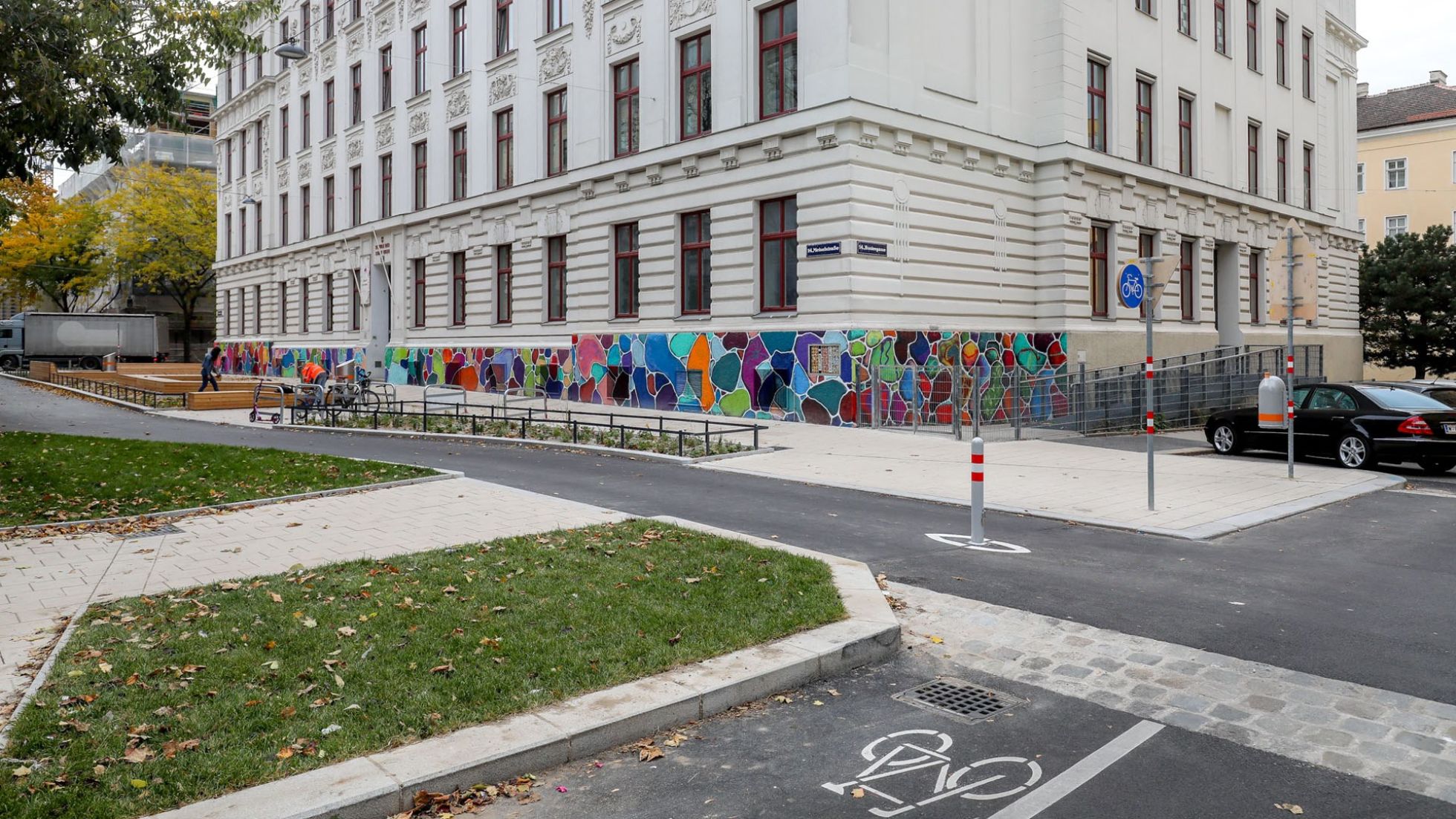 Kleine Straßenkreuzung; es kreuzen sich ein Gehweg, ein Fahrradweg und eine normale Fahrbahn; dahinter ein großes Gebäude mit bunter bemalung im unteren Bereich;